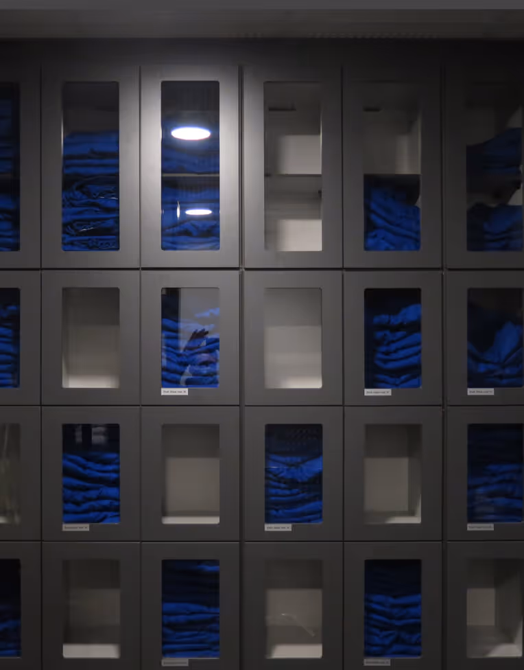Dark grey lockers with glass windows, some filled with neatly folded blue clothing and others empty.