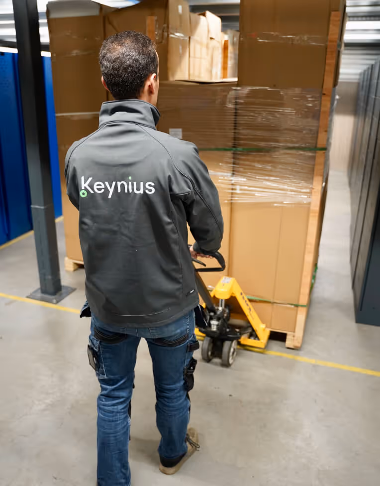 Worker in a Keynius jacket using a pallet jack to move a large wrapped pallet of boxes in a warehouse.