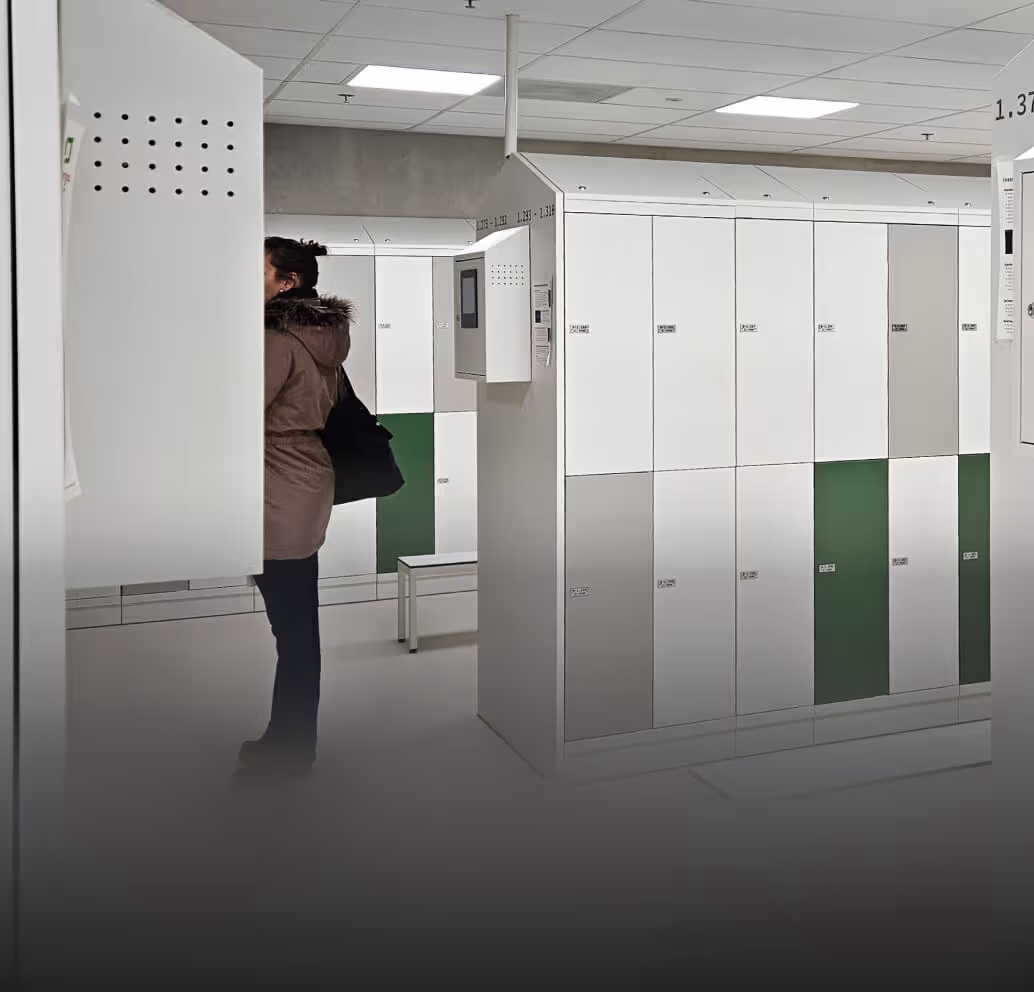 Person wearing a brown coat standing in a locker room with white and green lockers and benches.