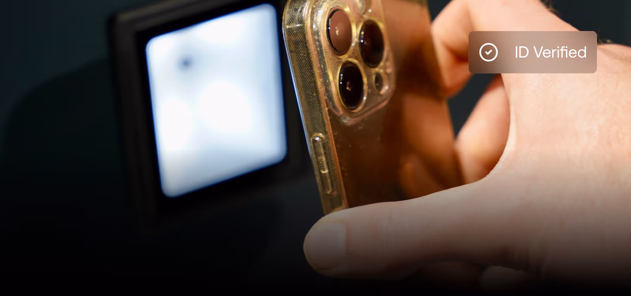 Hand holding a smartphone near a blurred screen with a notification showing a checkmark and the text 'ID Verified'.