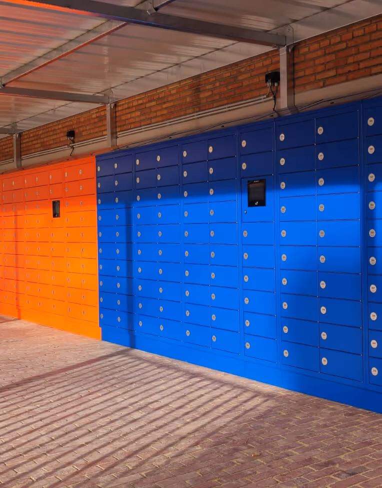 Rows of orange and blue parcel lockers under a metal roof with brick wall backdrop and long shadows cast on the brick floor.