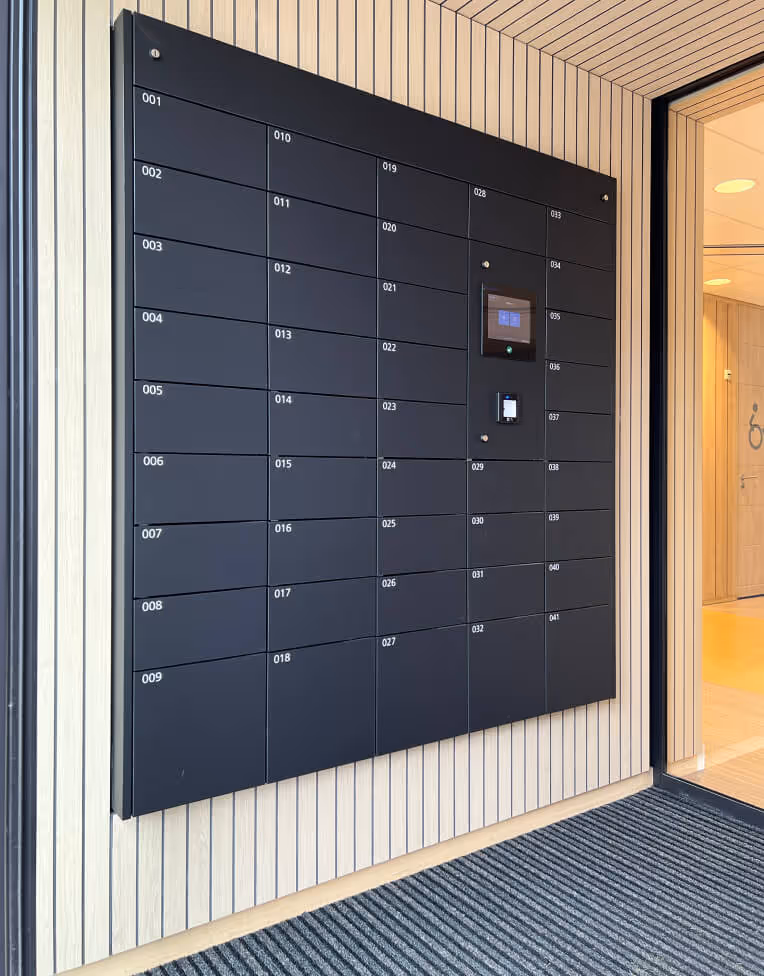 Black parcel lockers with numbered compartments mounted on a light wood-paneled wall next to a glass entrance door.