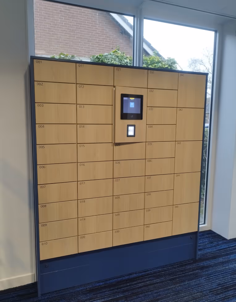 Wooden parcel lockers with numbered compartments and a digital touchscreen in an indoor space by large windows.