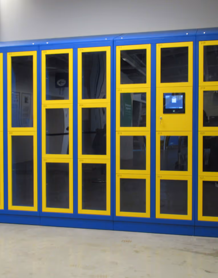 Wall of blue and yellow parcel lockers with glass doors and a digital screen on one locker.