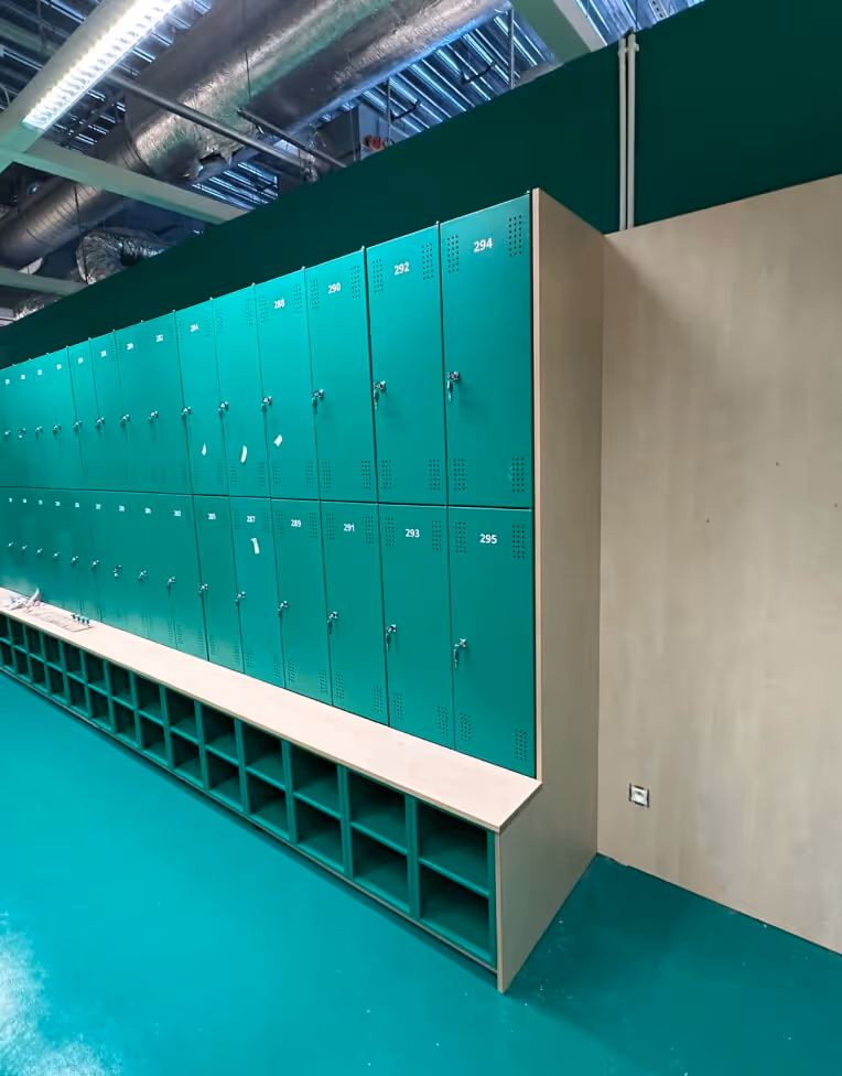 Row of teal lockers with numbered doors and open cubbies below in a clean, industrial-style room with exposed ceiling pipes.