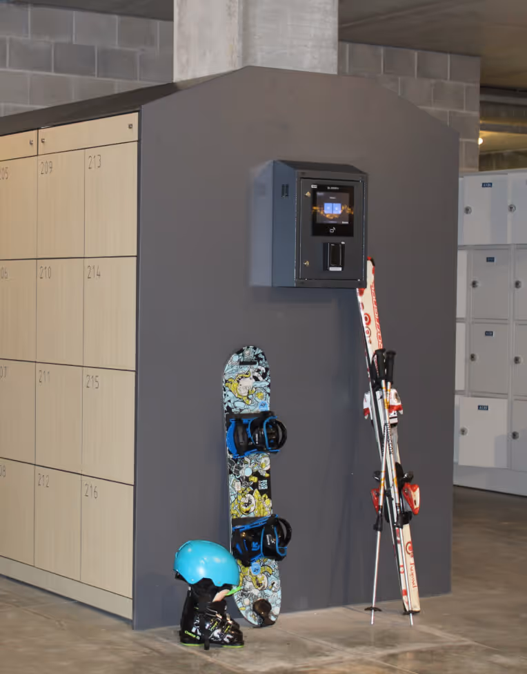 Snowboard, ski poles, skis, a blue helmet, and ski boots leaning against a gray wall with lockers nearby in an indoor setting.
