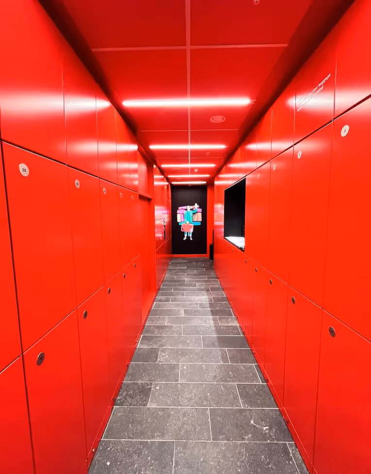 Narrow hallway with bright red lockers on both sides, gray tiled floor, and a colorful artwork at the far end.