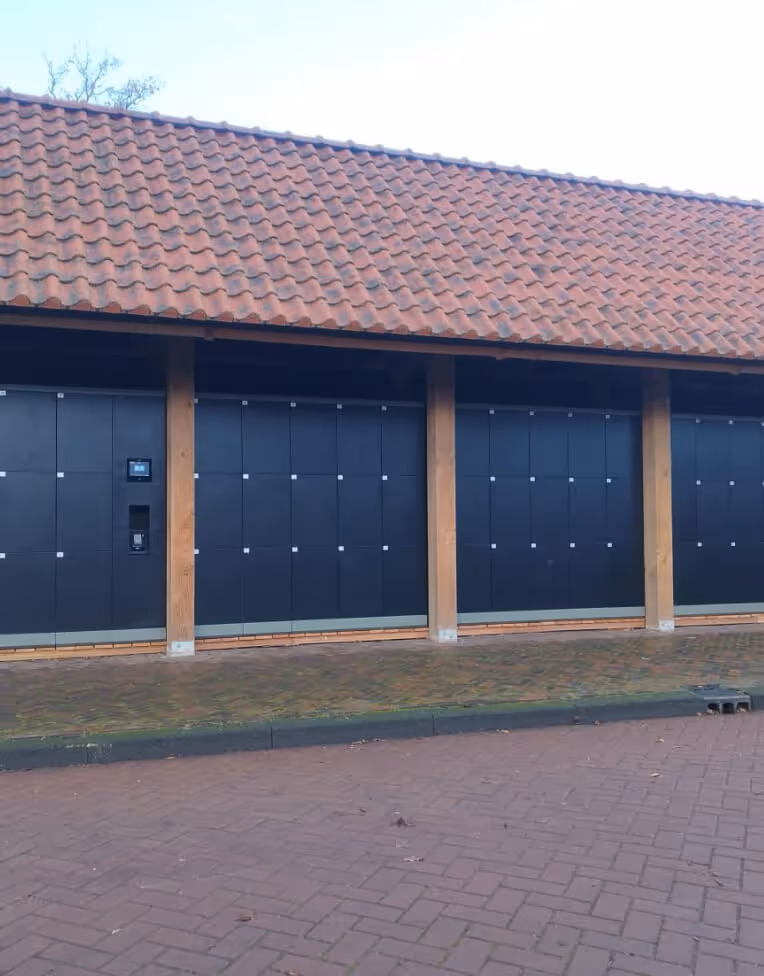 Row of black parcel lockers under a tiled roof with wooden pillars on a paved surface.