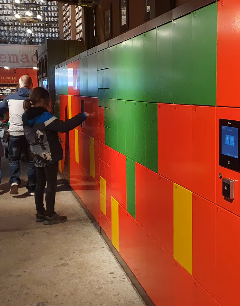 Person accessing a colorful self-service locker system with red, green, and yellow doors inside a commercial or public space.