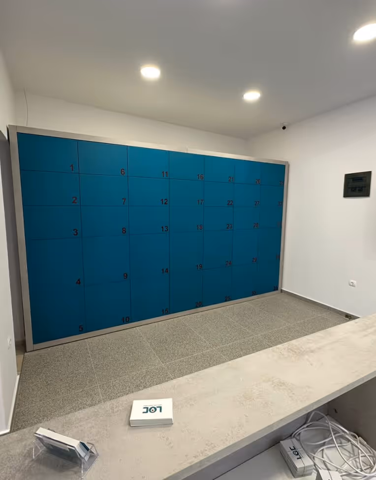 Interior room with a large blue wall-mounted locker unit numbered in rows, a grey tiled floor, and a beige countertop with some small boxes and cables on it.