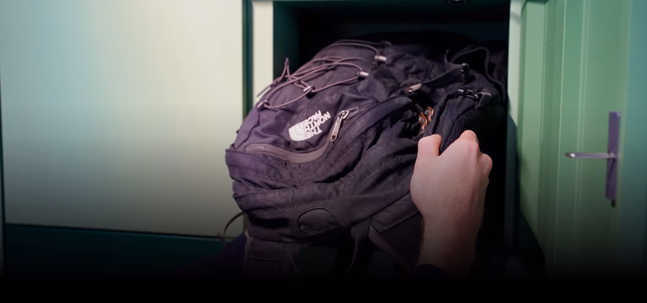 Hand placing a black North Face backpack into a green locker.