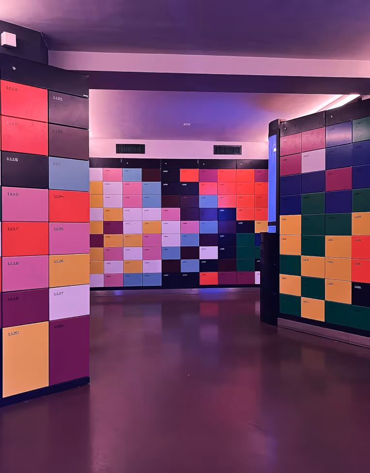 Indoor room with walls covered in multiple colorful square lockers with small label tags, under soft purple and pink lighting.