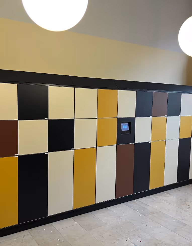 Wall of multicolored lockers in beige, black, brown, and mustard yellow with a digital screen panel in the middle.
