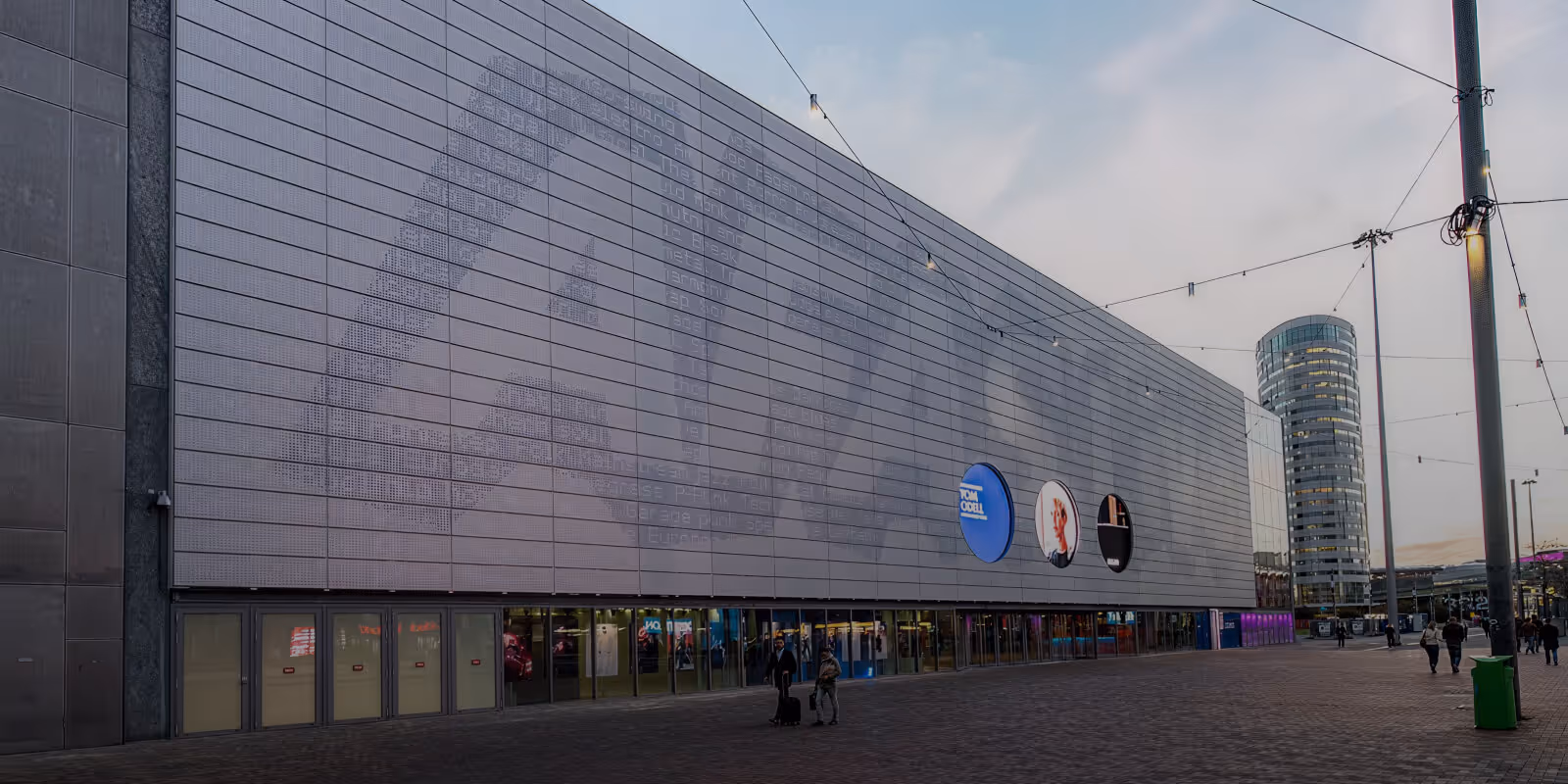 Modern building with a large patterned facade and three circular displays, located in an open plaza at dusk.