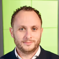 Portrait of a man with short curly hair and a trimmed beard, wearing a dark sweater over a white collared shirt, against a green background.
