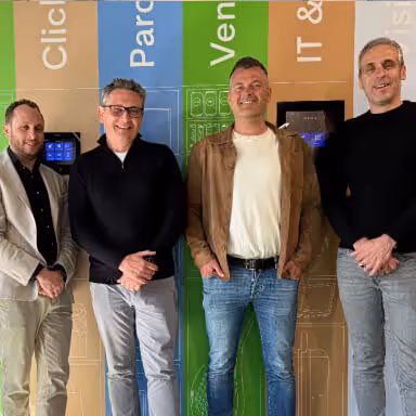 Four men standing indoors in front of a colorful wall with technical drawings and text, smiling at the camera.