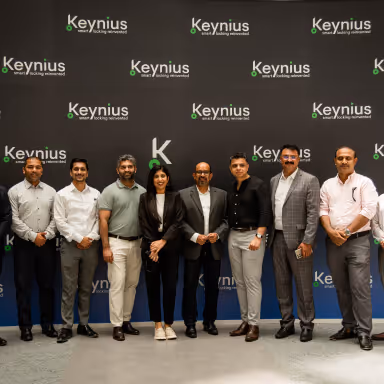 Group of eight professionally dressed people standing in front of a Keynius branded backdrop.