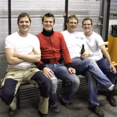 Four young men sitting together on a bench in casual clothing, smiling at the camera, one holding a bottle.