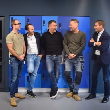 Five men casually talking and smiling in front of blue lockers, with some wearing jeans and casual tops and one in a suit.