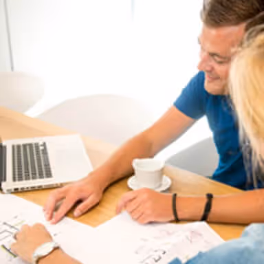 Two people working together at a table with a laptop and papers with sketches and charts.