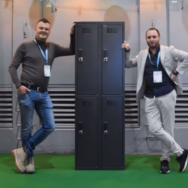 Two men standing on either side of a tall black locker unit on a green floor, both wearing conference badges.