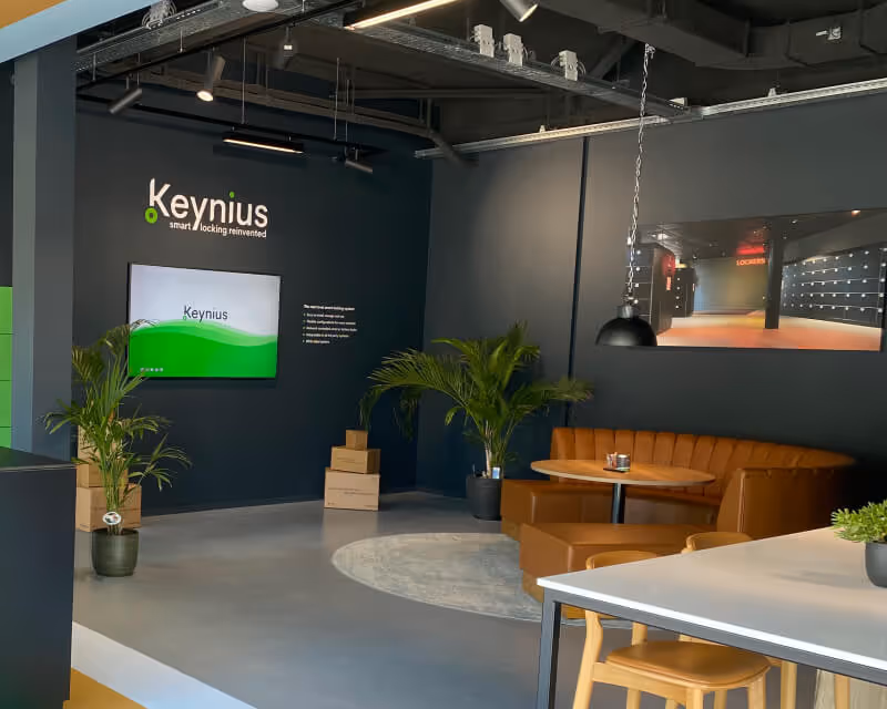 Modern office corner with dark walls featuring Keynius branding, a mounted screen, potted plants, stacked boxes, a round table with brown leather seating, and a hanging industrial light.