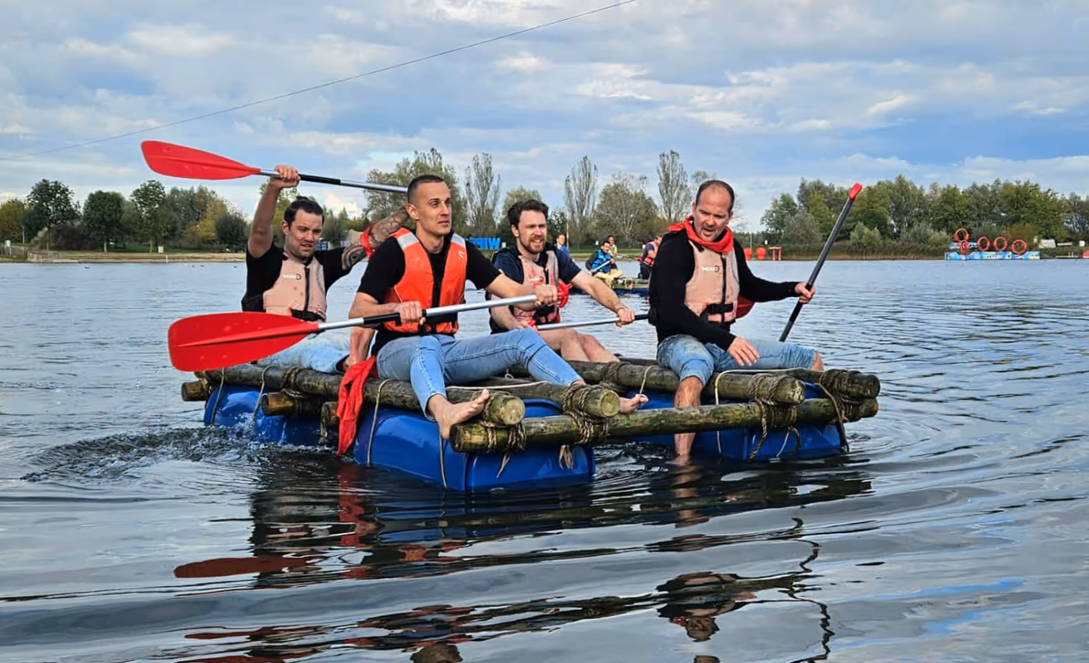 Four men wearing life jackets paddling a makeshift raft with wooden logs and blue barrels on a lake under a cloudy sky.