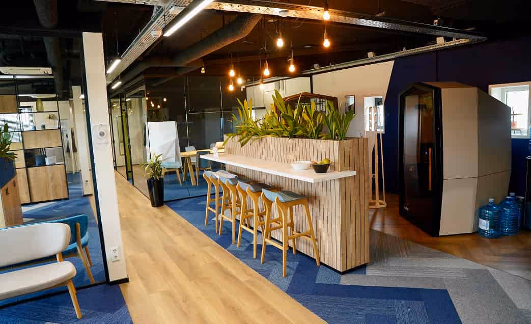 Modern office lounge area with wooden bar counter, high stools, indoor plants, blue carpet, and a whiteboard in background.