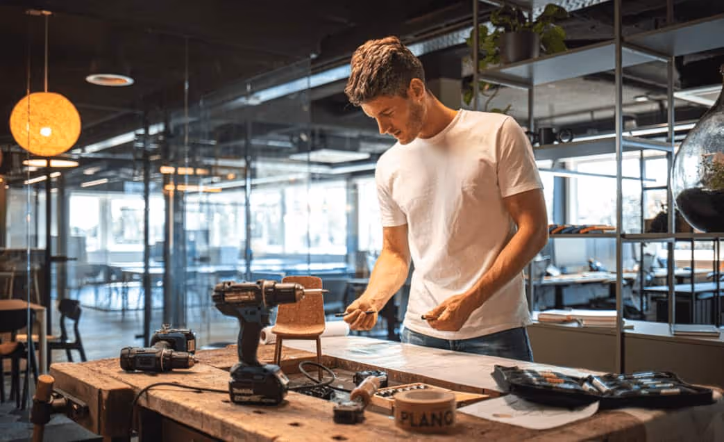 Man in a white t-shirt working on woodworking project at a table with tools in a modern workshop.