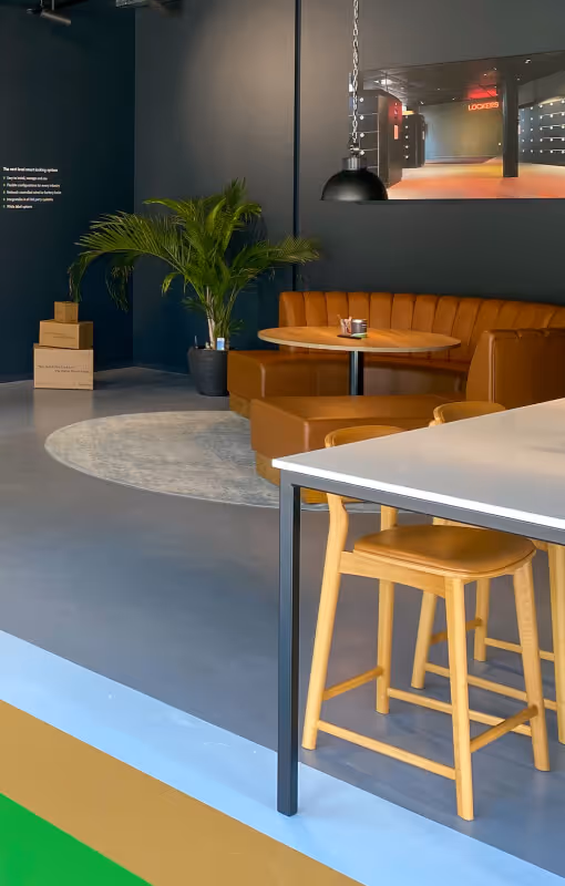 Modern office lounge area with a round wooden table, tan leather L-shaped sofa, two wooden chairs, a potted palm plant, and a large wall photo of an interior space.