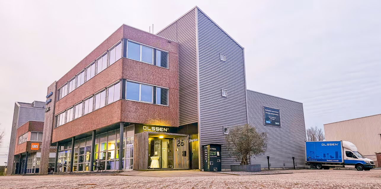 Modern commercial building with brick and metal siding, featuring glass windows, an entrance with 'OLSSEN' signage, and a blue delivery truck parked outside.