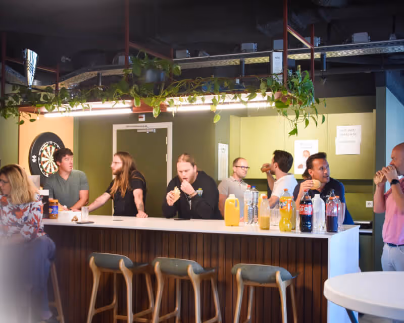 Group of people standing and sitting around a modern bar counter with drinks and snacks, engaged in conversation and eating.