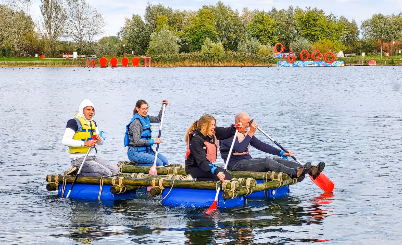 Four people paddling on a homemade raft made of wooden logs and blue barrels on a calm lake with green trees in the background.