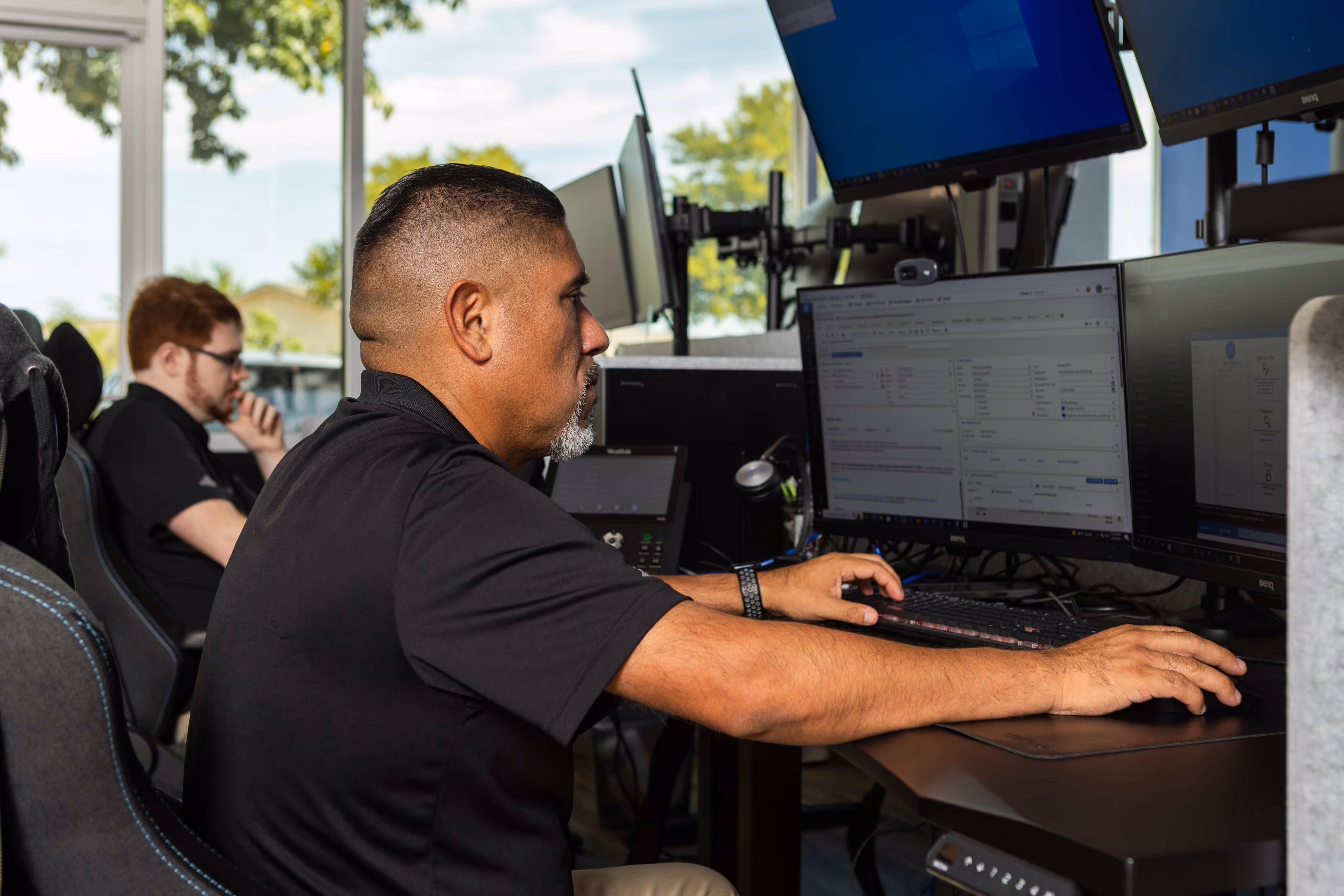 IT technician working at multi-monitor workstation in Austin TX managed services office with colleague in background