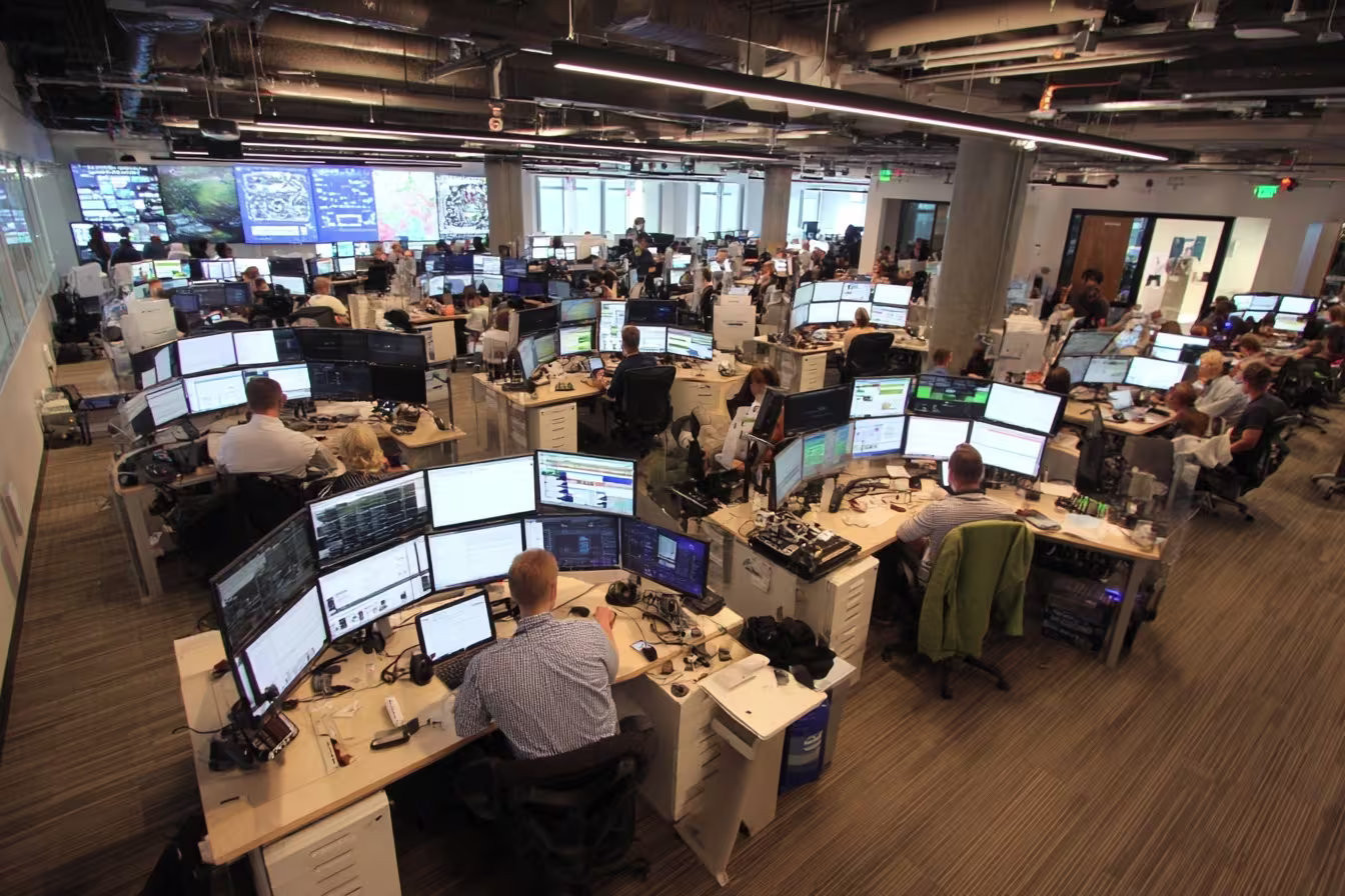 Large open-plan IT operations center with technicians monitoring multiple screens and cybersecurity dashboards
