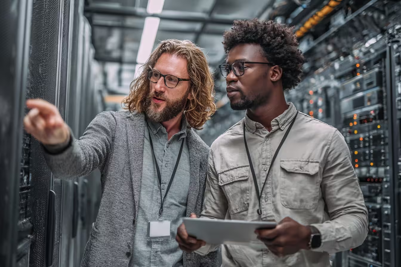 Two IT technicians reviewing server rack equipment in a data center, one pointing while the other holds a tablet
