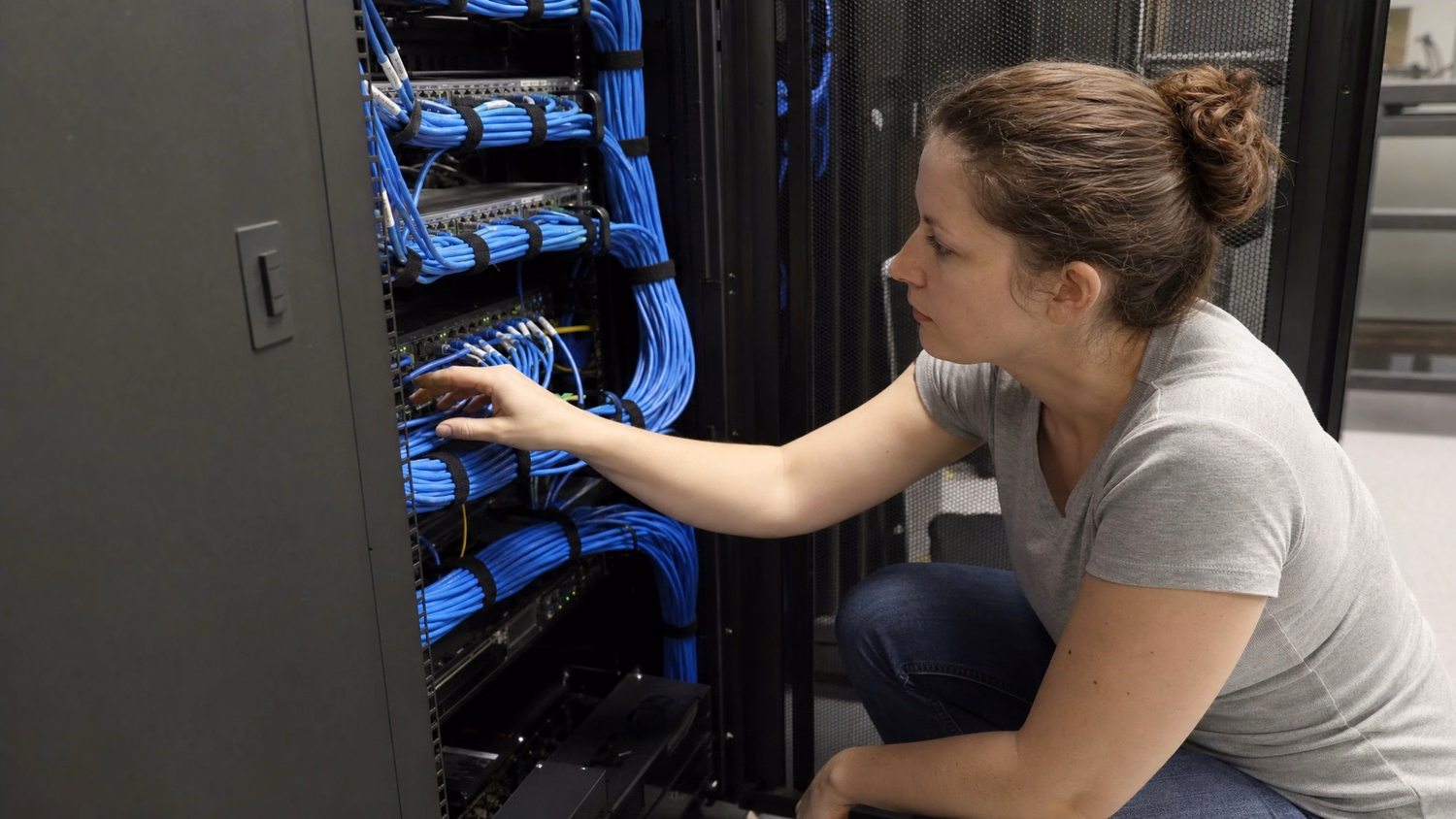 A white woman in a network closet crouches, tracing a cable run on a patch
