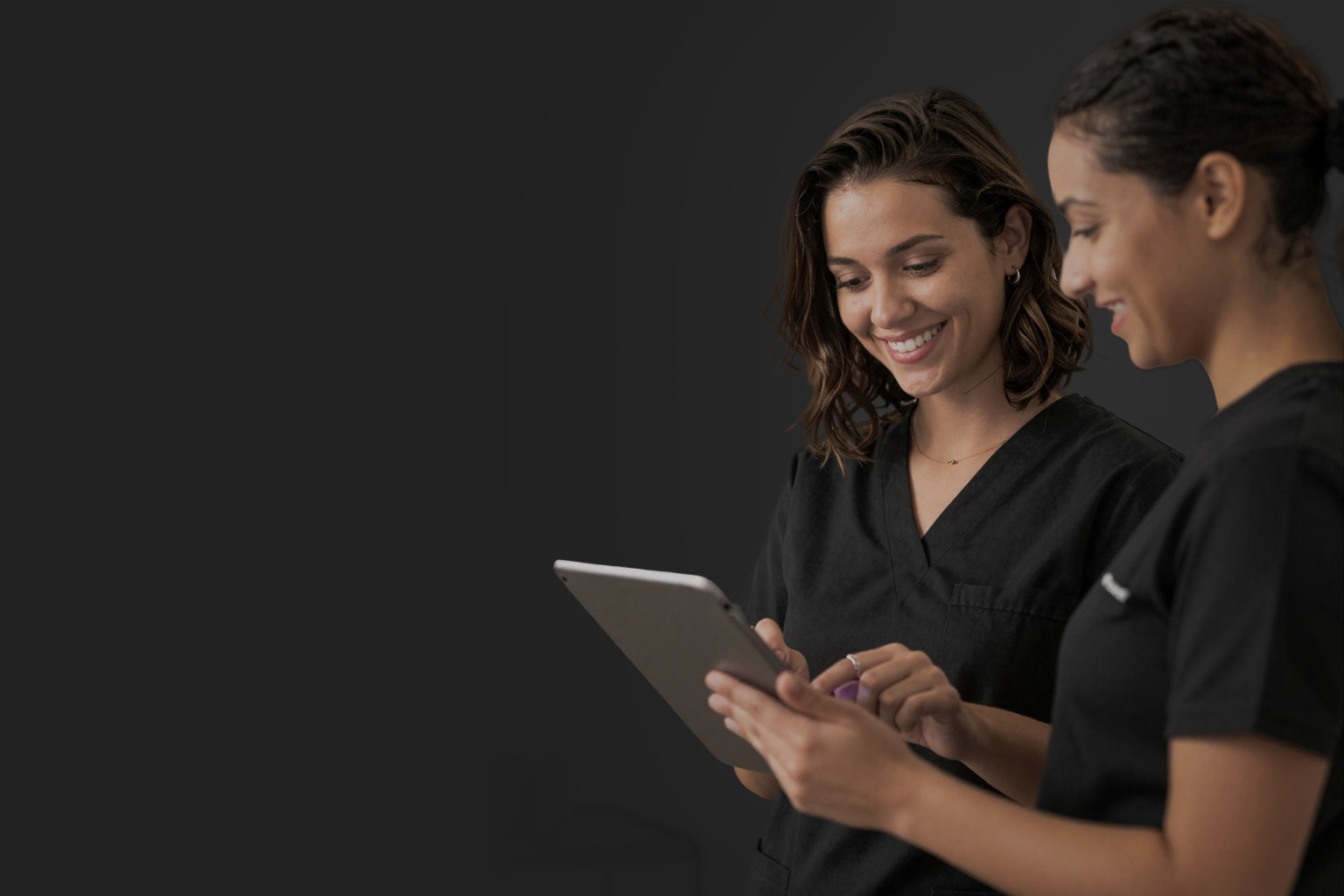Duas mulheres sorrindo usando uniformes pretos enquanto olham para um tablet.