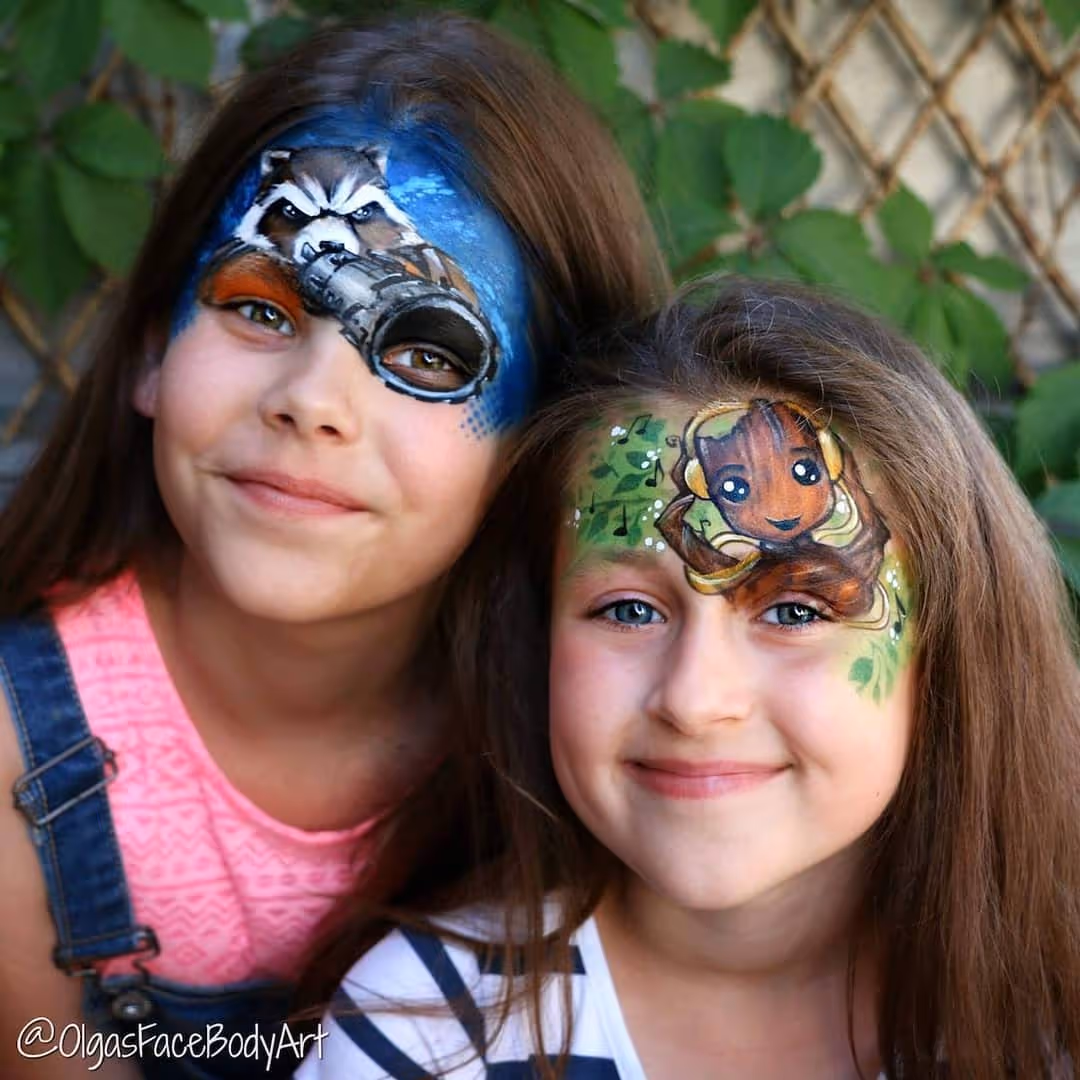 Two smiling girls with face paint: one has a raccoon and scope, the other has a Baby Groot design.