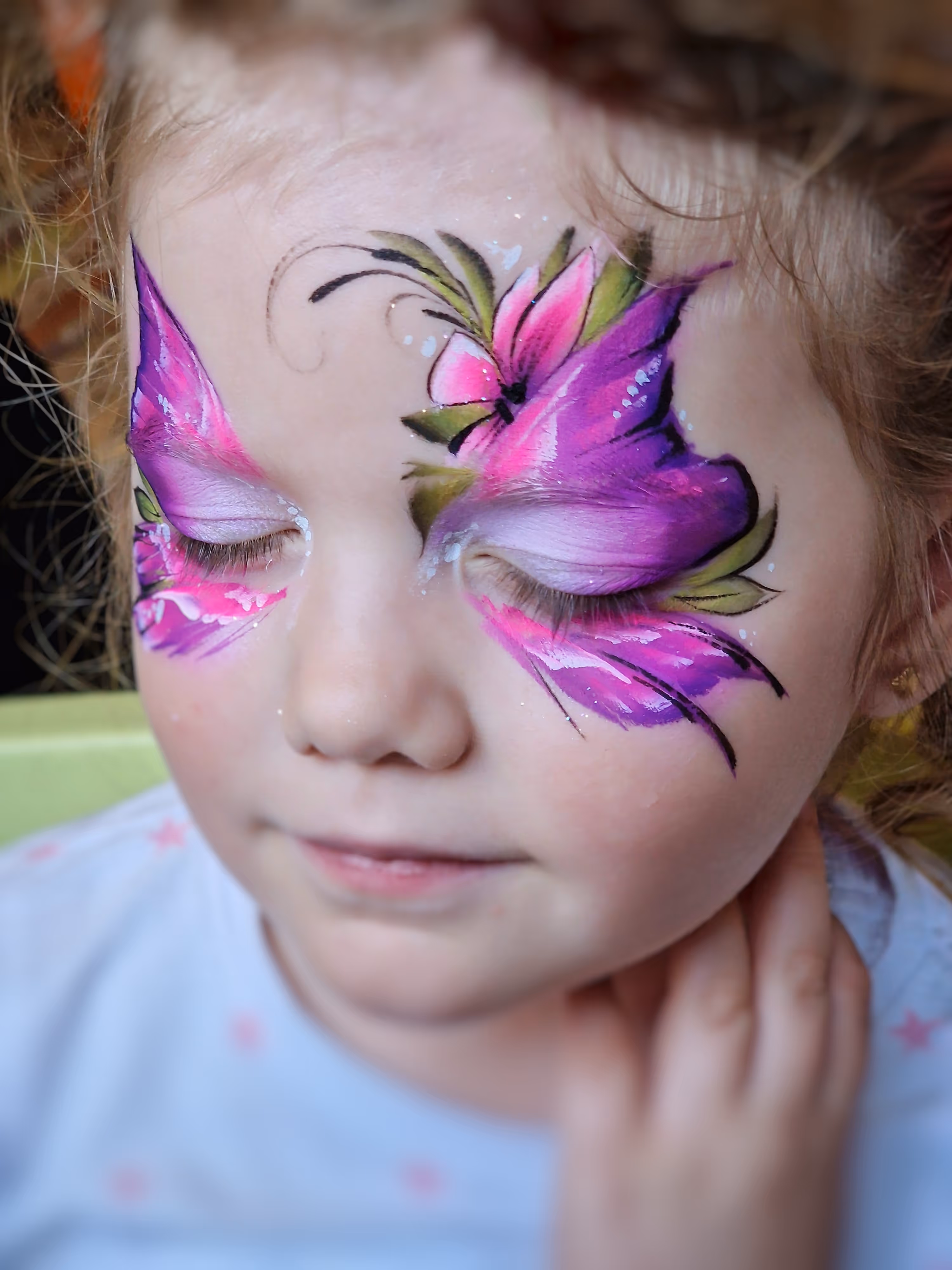 Young girl with closed eyes and pink-purple butterfly face paint with green leaf accents.