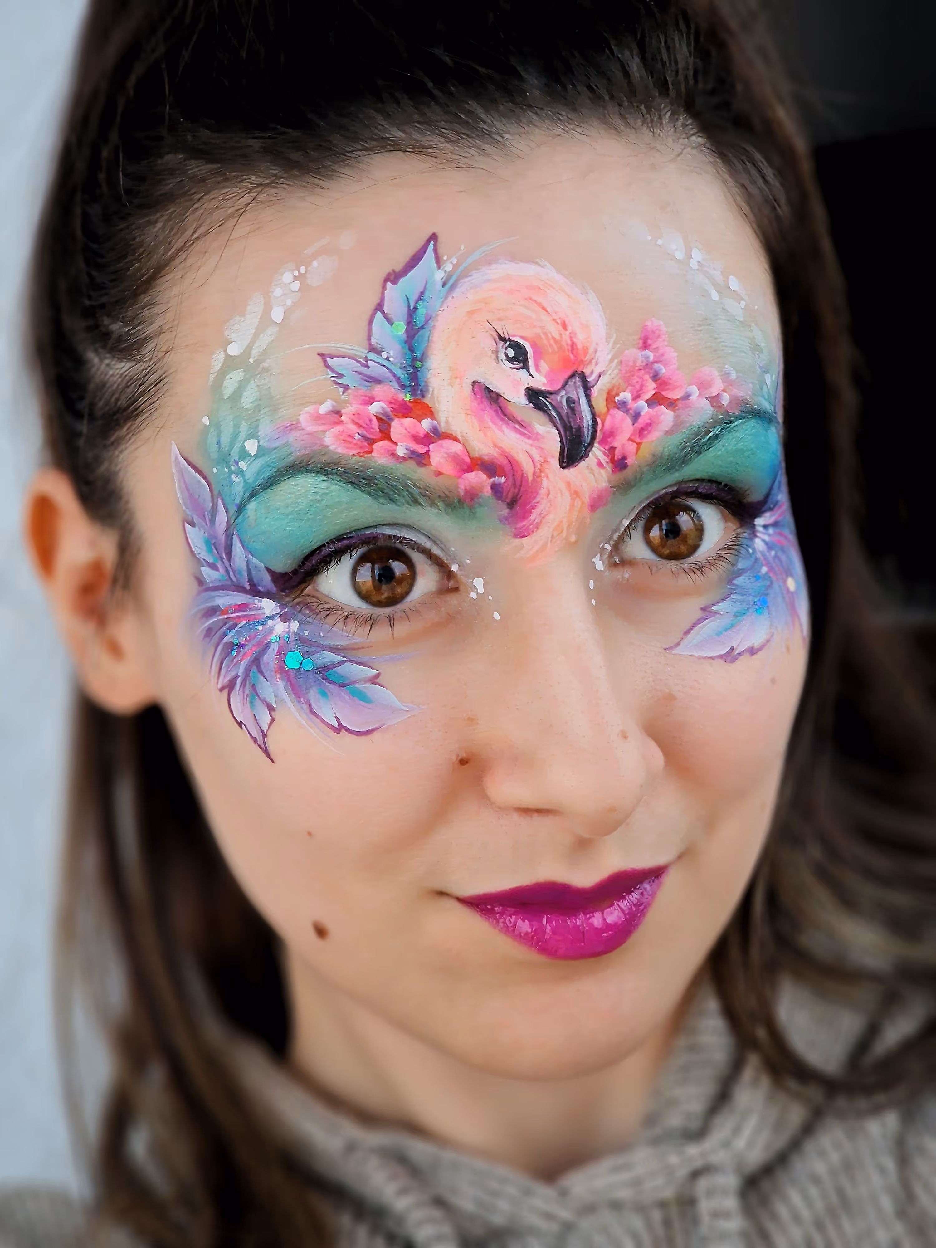 Woman with face paint of a pink flamingo surrounded by colorful flowers and leaves around her eyes.