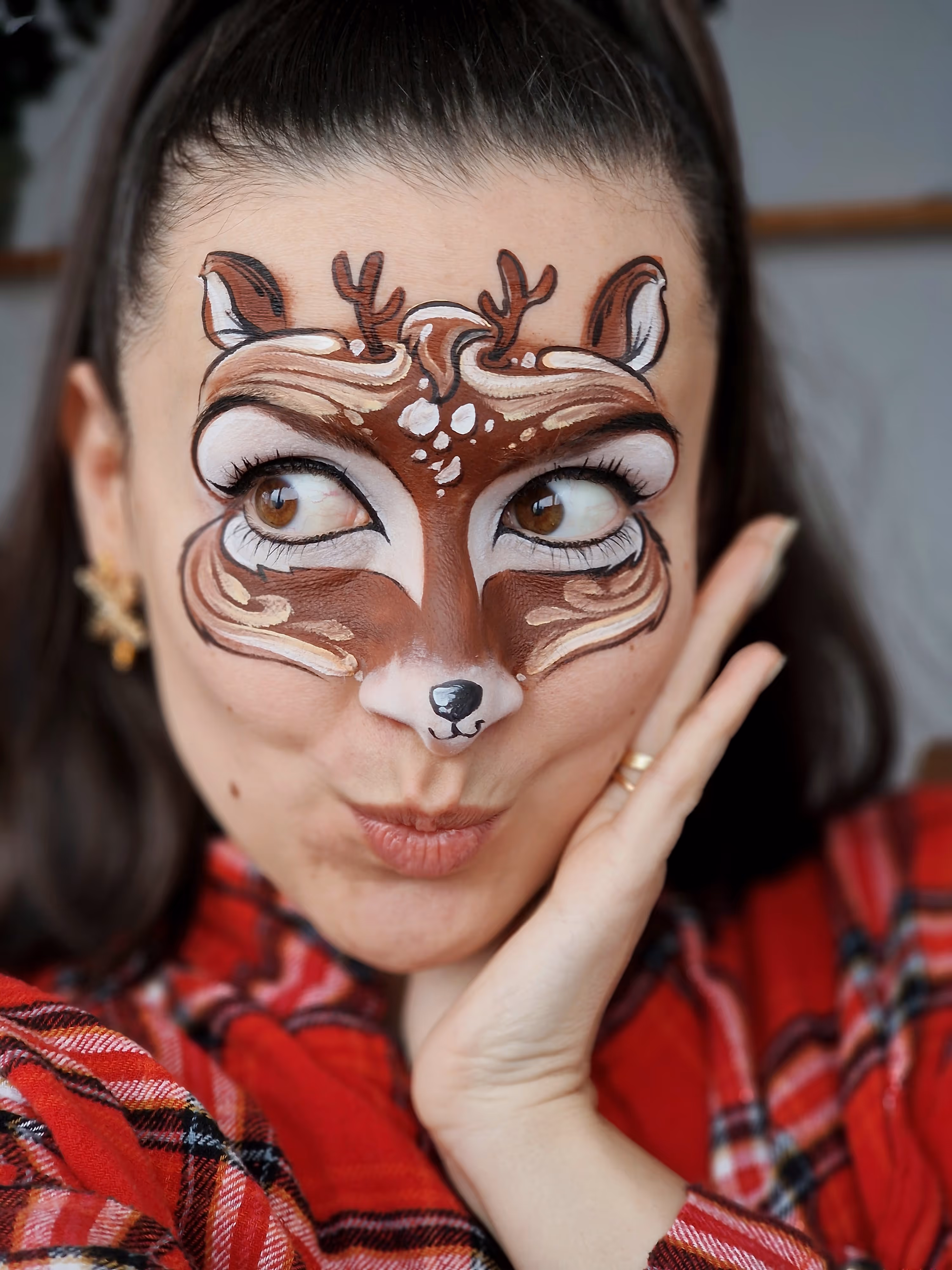 Woman with face paint of a brown deer with antlers and ears wearing a red plaid shirt.
