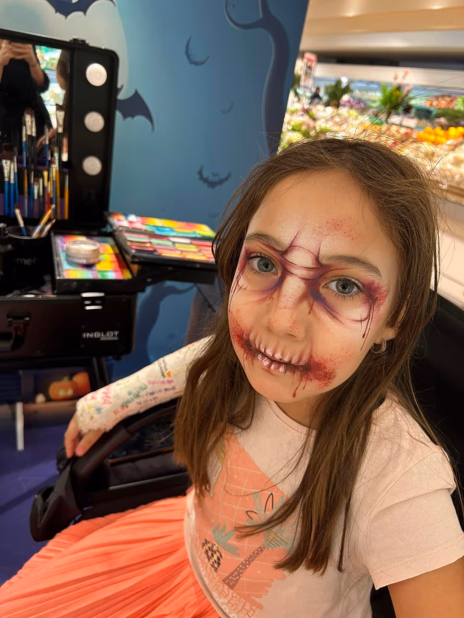 Young girl with Halloween face paint resembling stitched scars and blood, sitting with colorful makeup kit nearby.