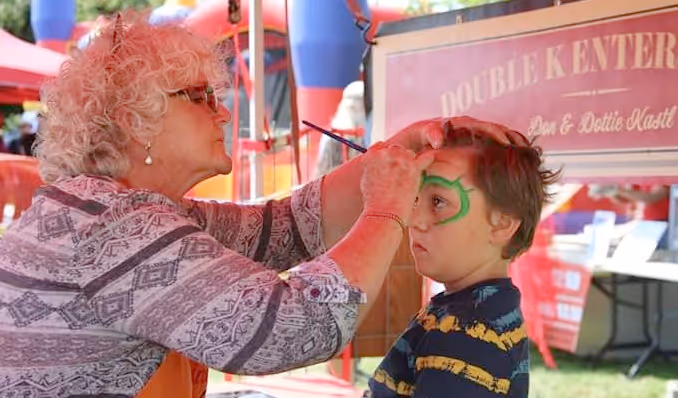 Woman painting green face paint on a young boy's face at an outdoor event.