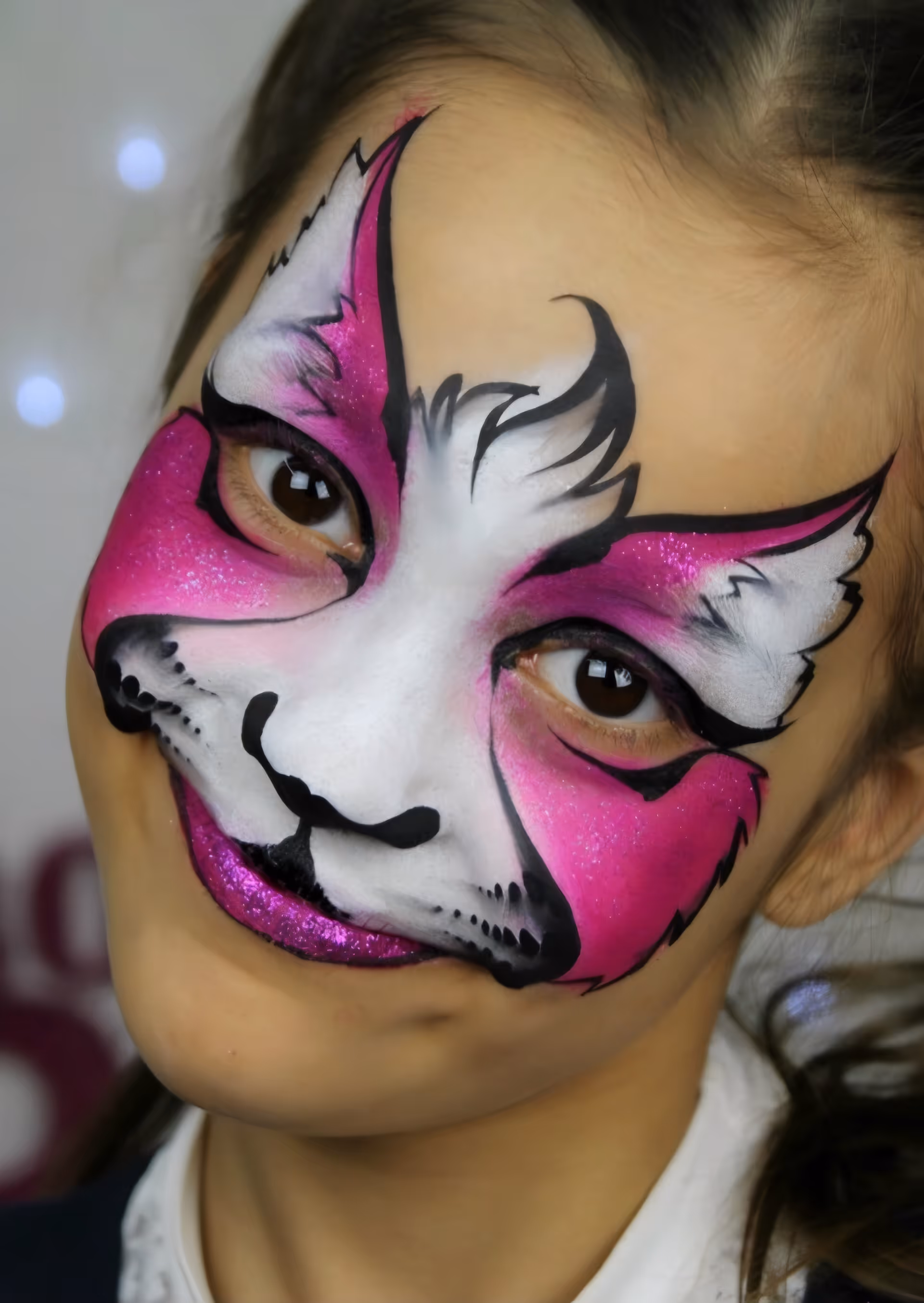 Face painted like a pink and white cat with black details, glitter lips, and expressive eyes.
