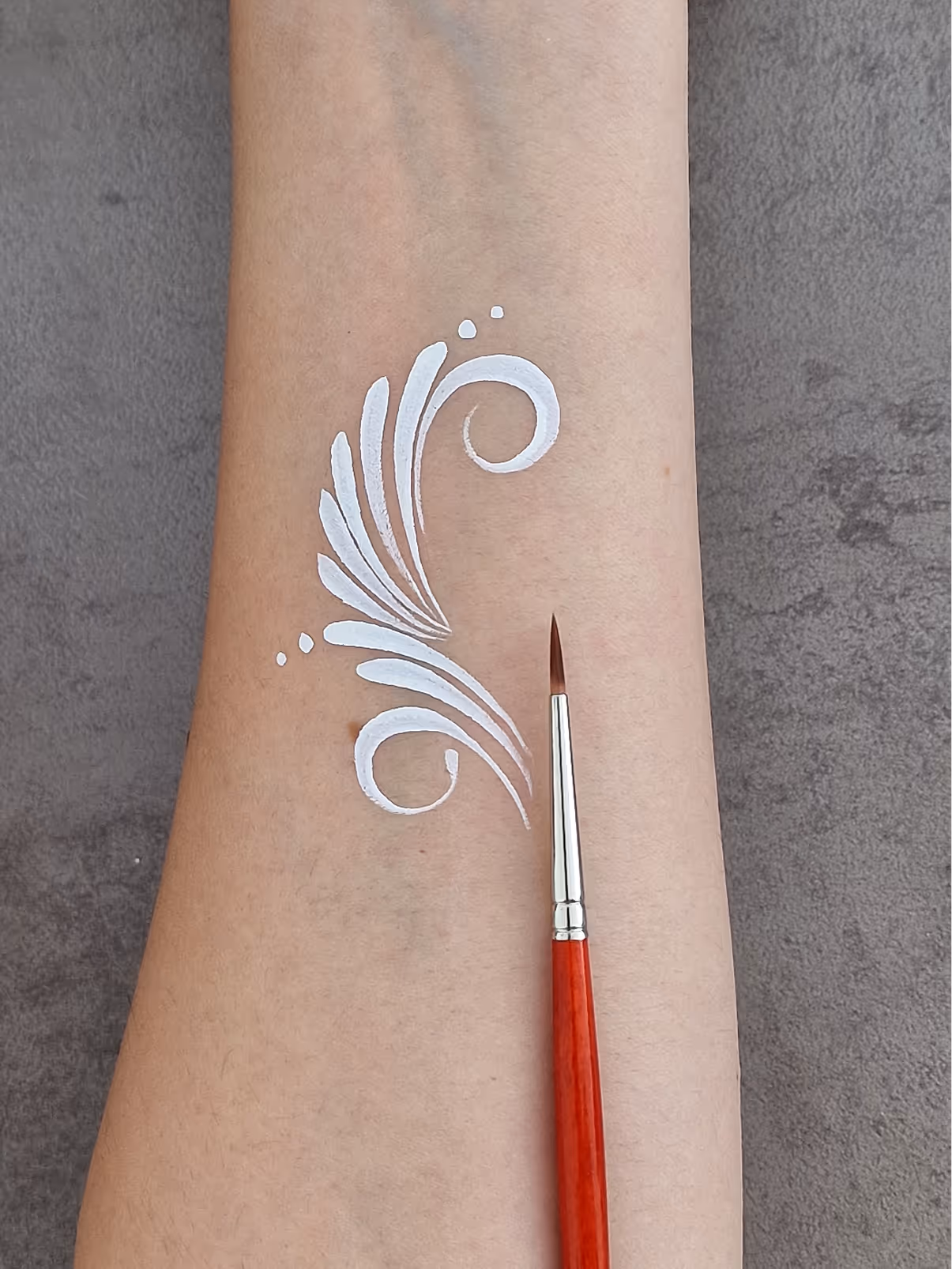White ornamental brush strokes painted on forearm with fine red-handled paintbrush.
