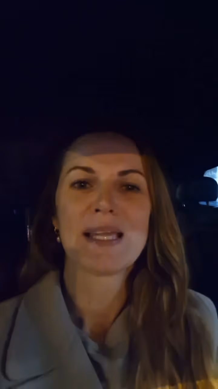 Woman with light brown hair speaking inside a car at night, wearing earrings and a coat.