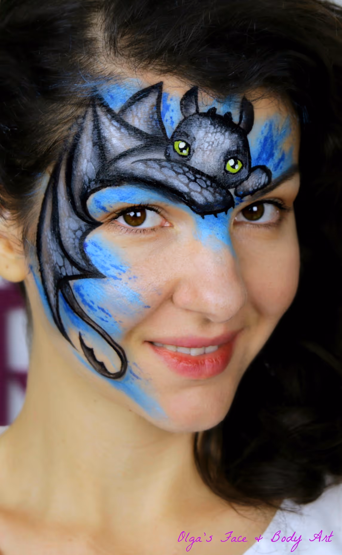 Woman smiling with face paint of a black dragon with green eyes on a blue background.
