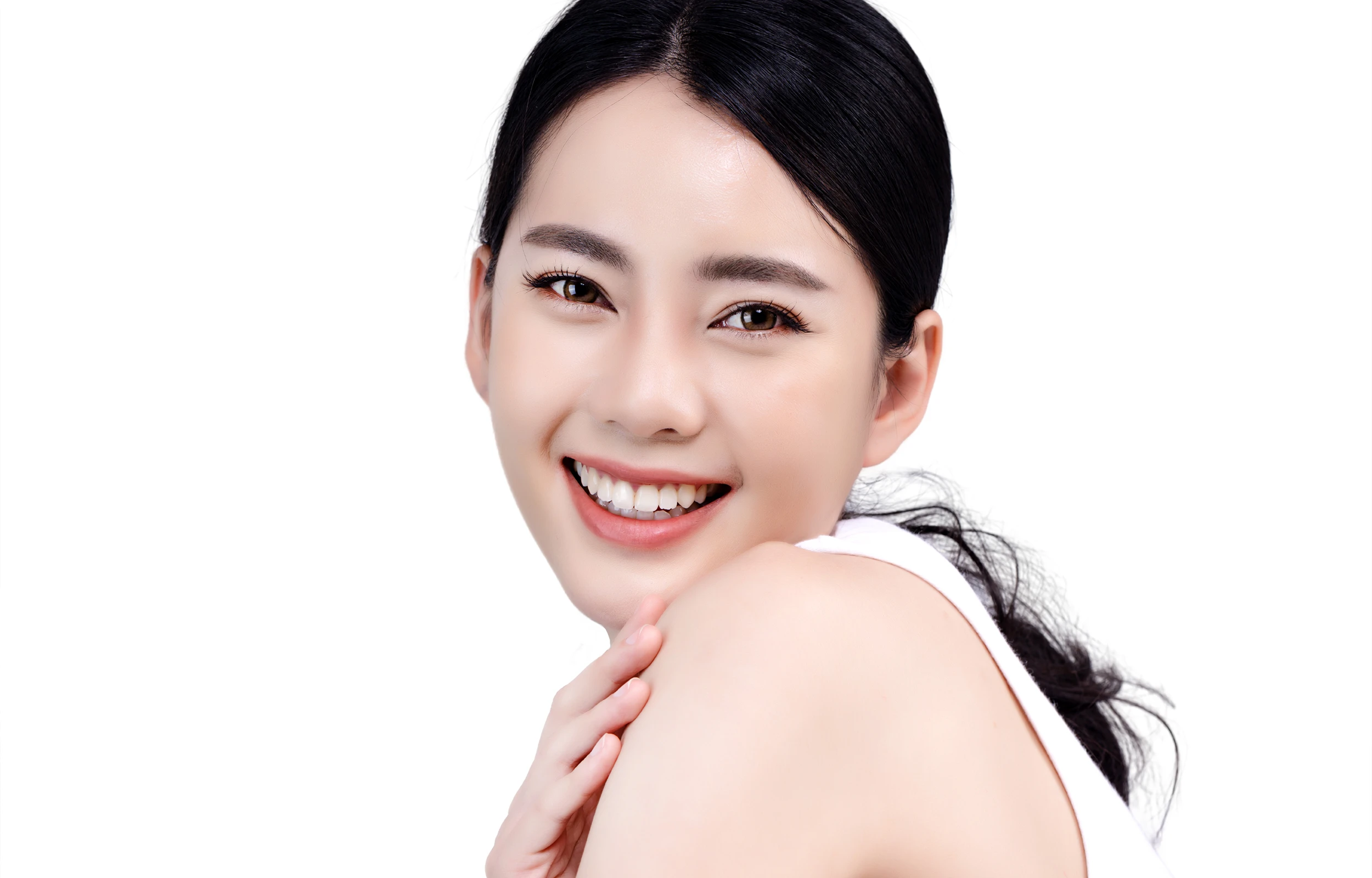 Close-up portrait of a smiling young woman with dark hair and clear skin, resting her hand on her shoulder against a white background.