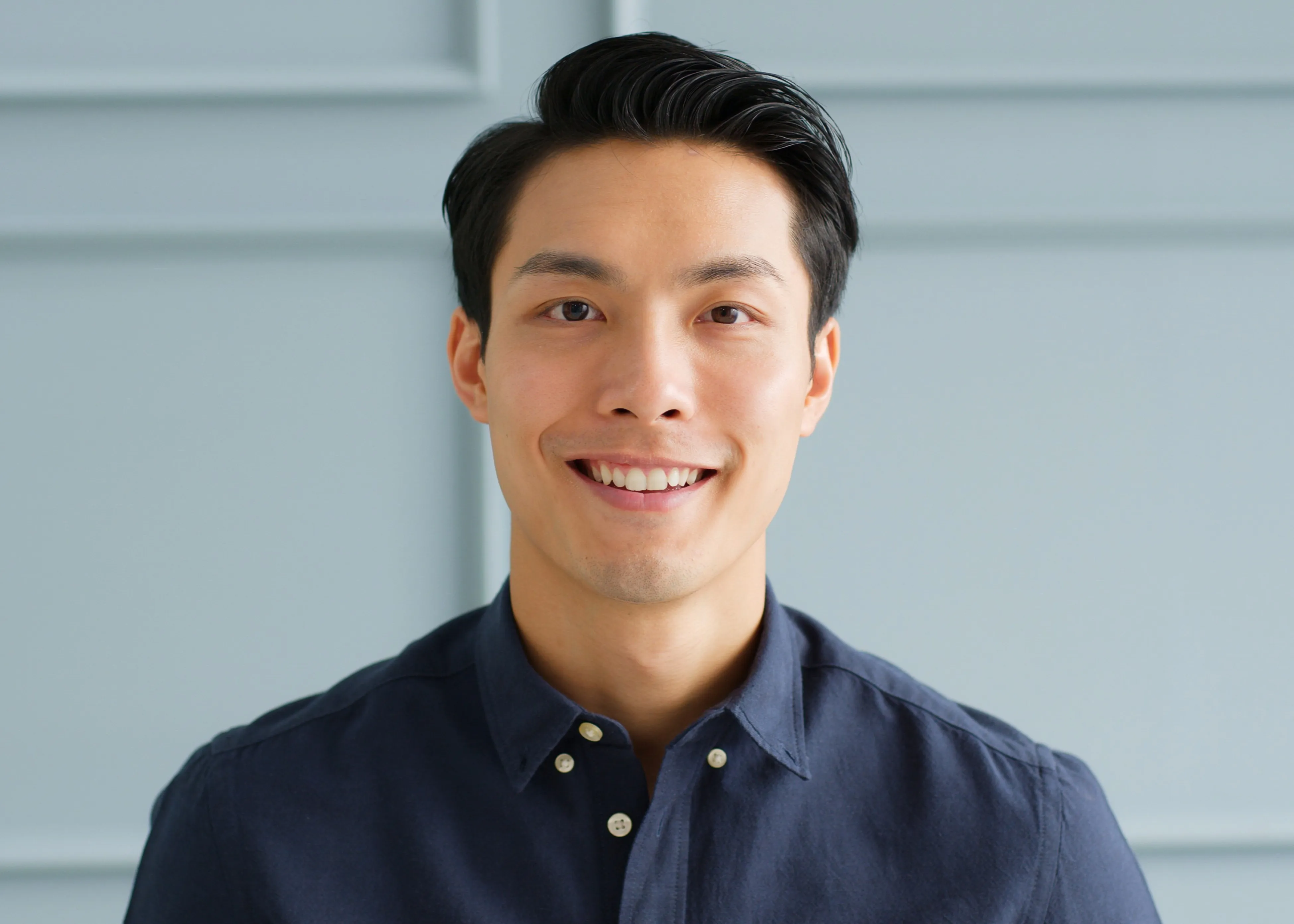 Smiling young Asian man with short black hair wearing a dark blue shirt against a light blue paneled wall.